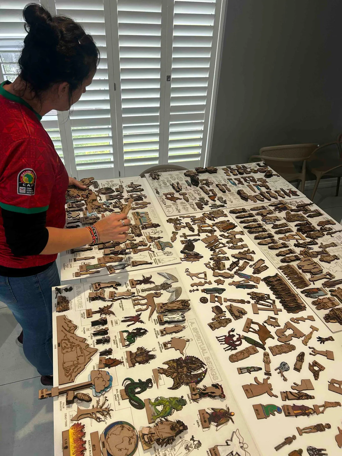 A woman in a red top sorting many small wooden figures laid out on a large white table.
