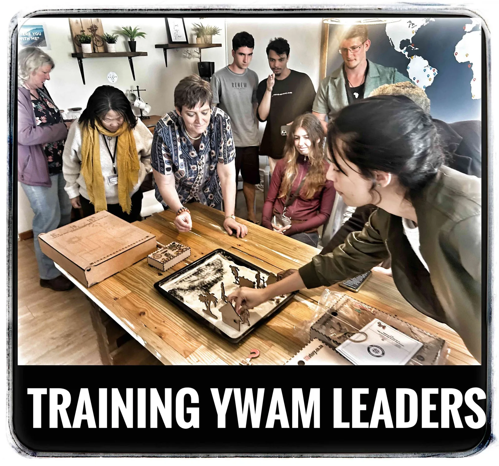 A group of people gathered around a table looking at wooden figures in a box lid, captioned Training YWAM leaders.